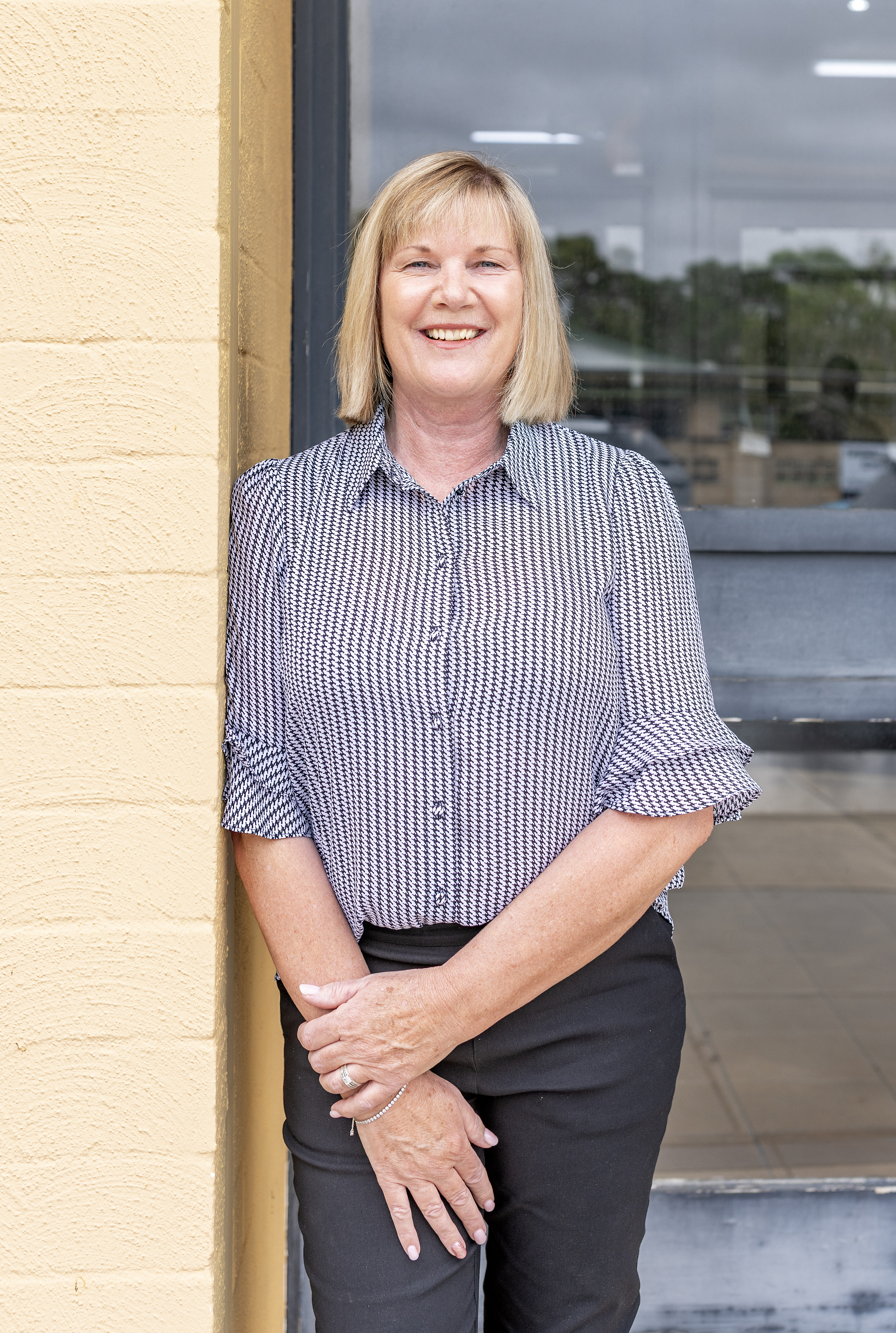 A middle-aged woman with blonde hair, dressed in a patterned shirt and black pants, stands in front of a glass door, her h...